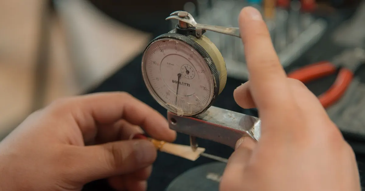 Hands holding a thin metal part under a dial indicator gauge to read a dimension, one of the tools used in CMM inspection of machined parts